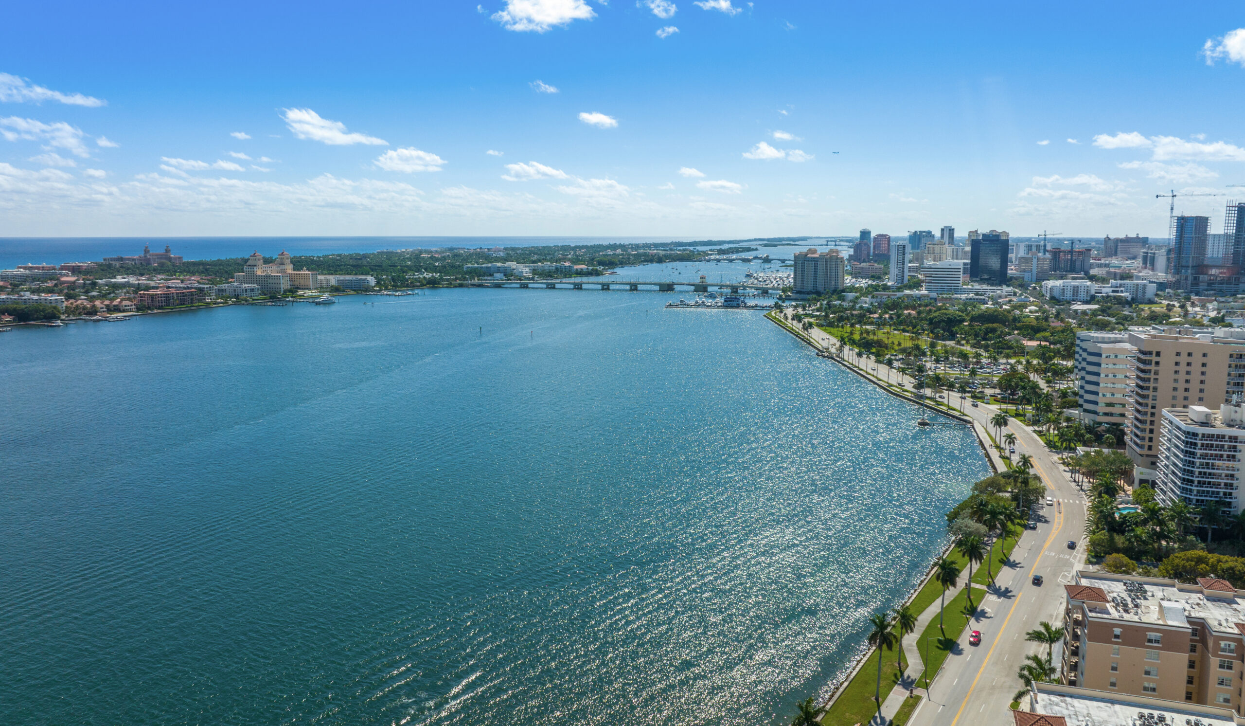 Aerial view of a coastal city skyline and waterfront with palm-lined boulevard, marina, and expansive ocean horizon.