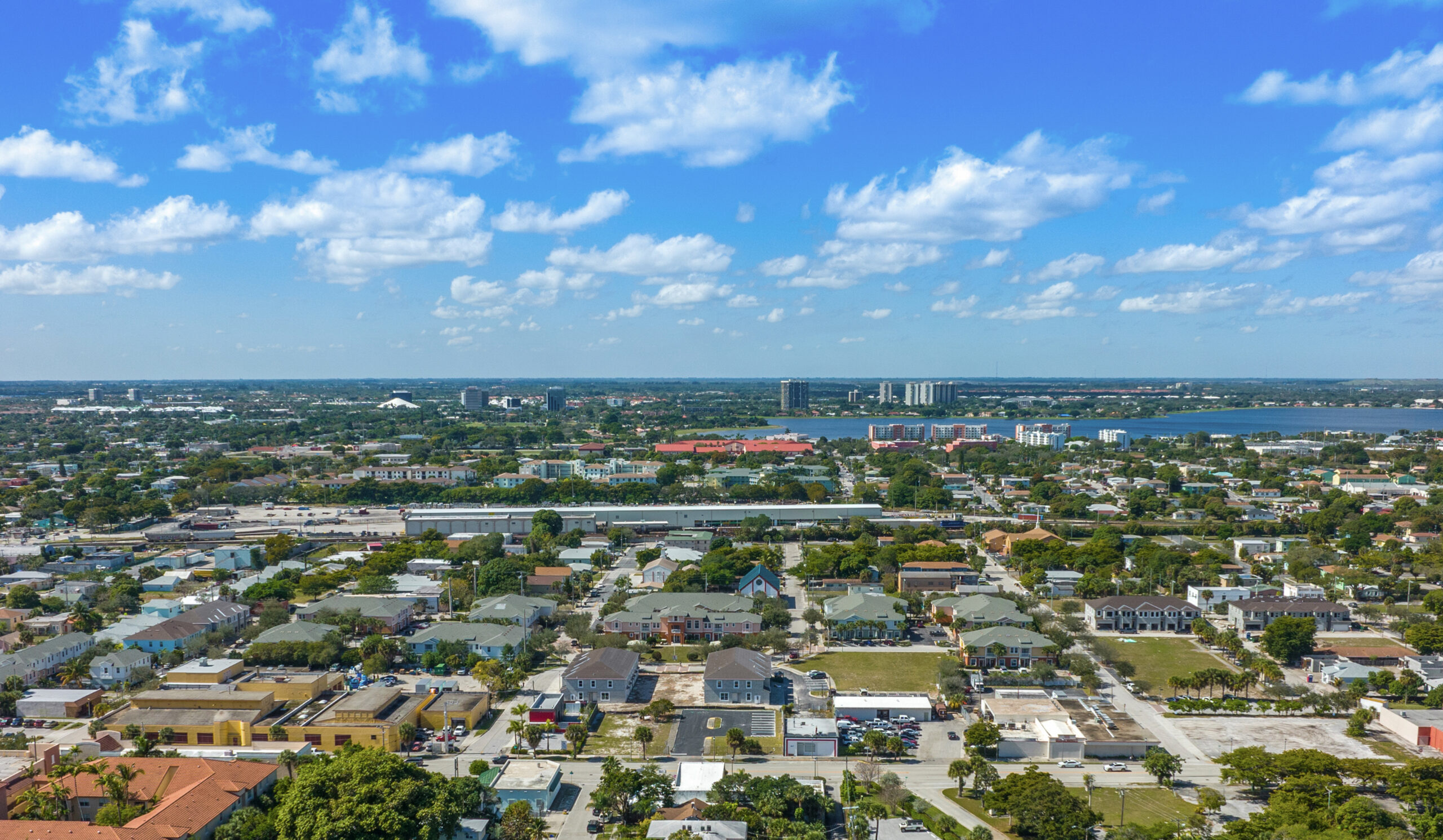 Aerial view of a coastal city neighborhood with low-rise buildings, tree-lined streets, and water visible in the distance.