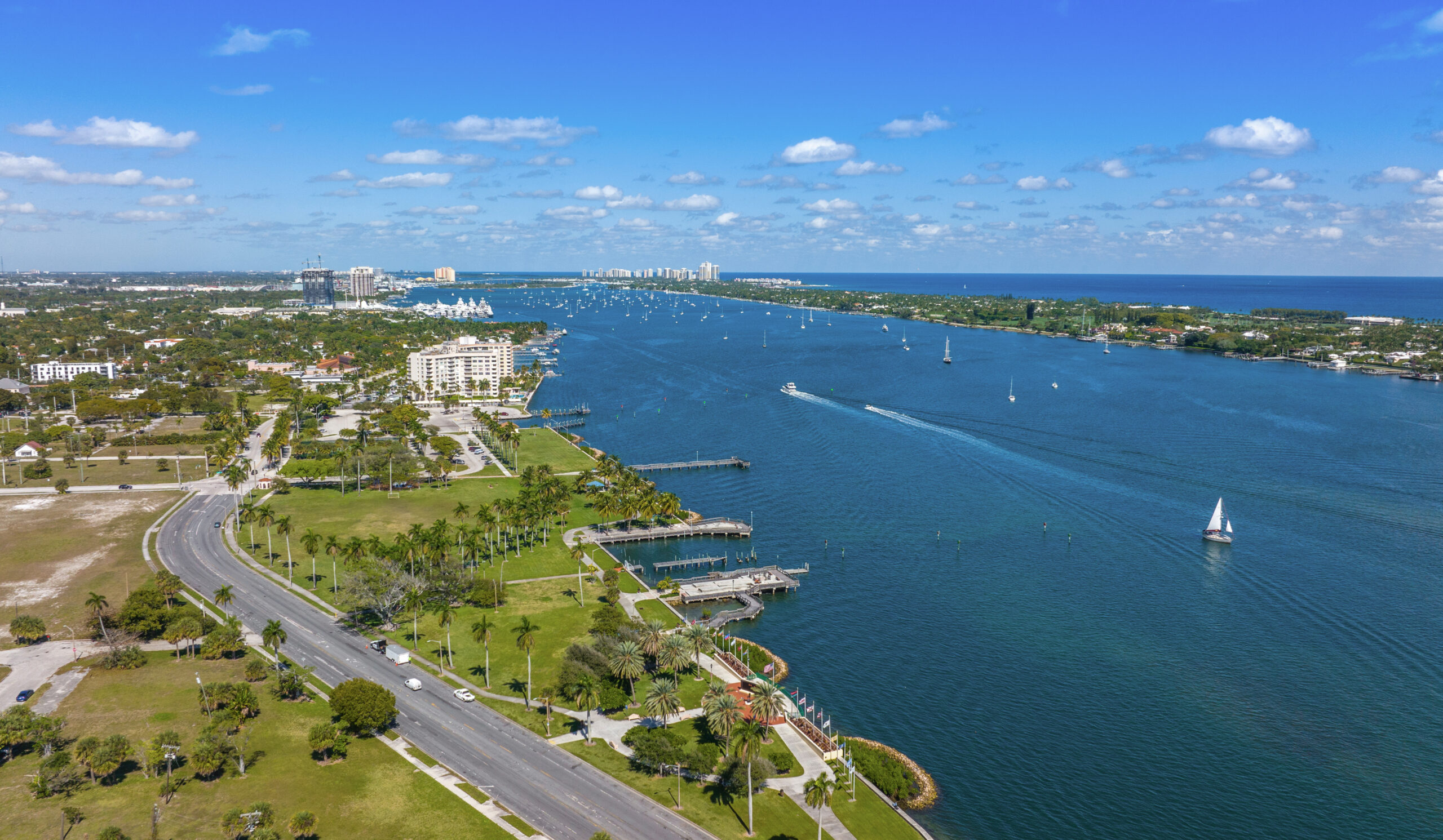 Aerial view of a coastal city waterfront with palm-lined park, marina docks, sailboats, and the ocean beyond.
