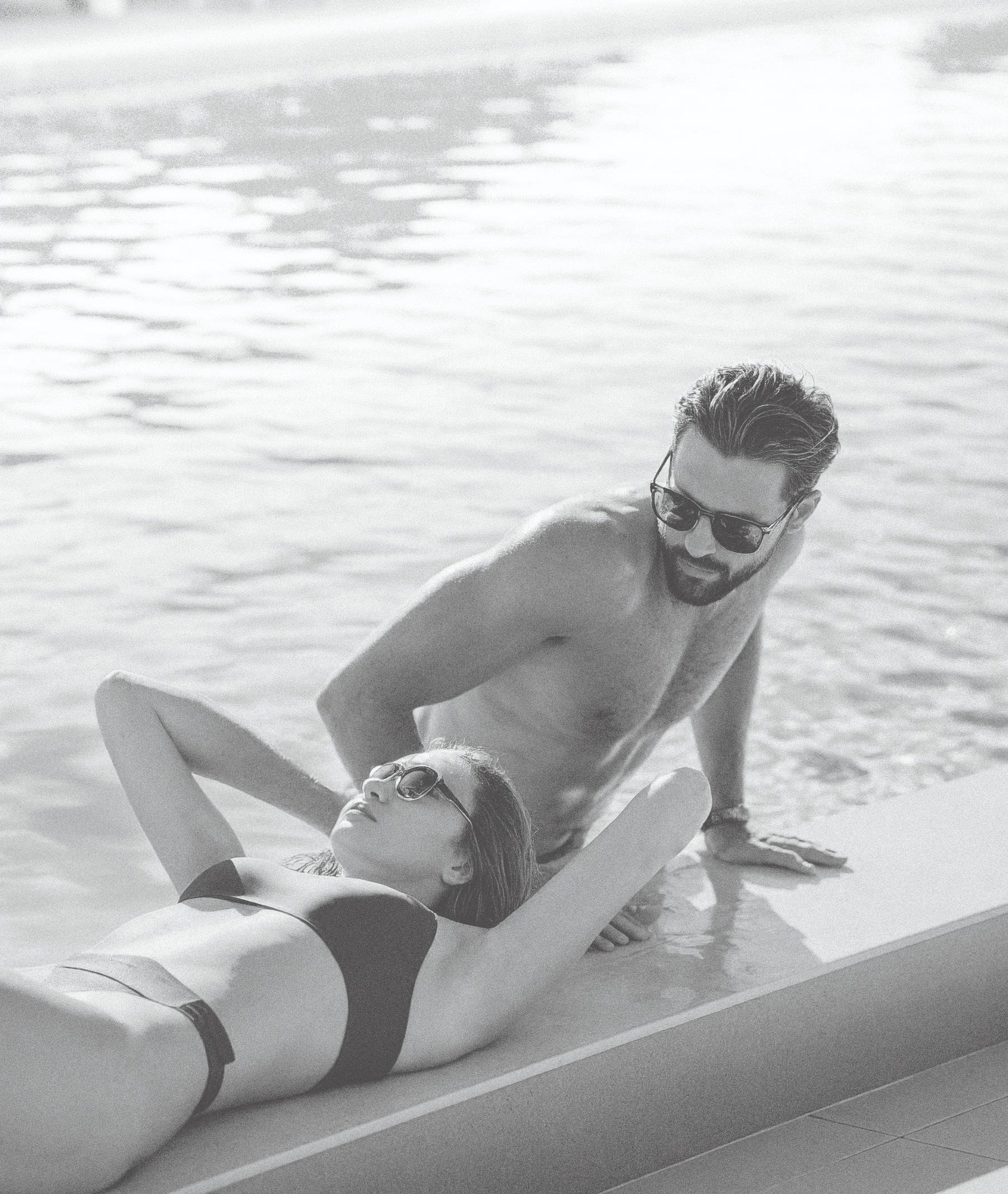 Black-and-white photo of a couple relaxing by a pool, with a woman in a bikini reclining at the water’s edge and a man wearing sunglasses leaning beside her.